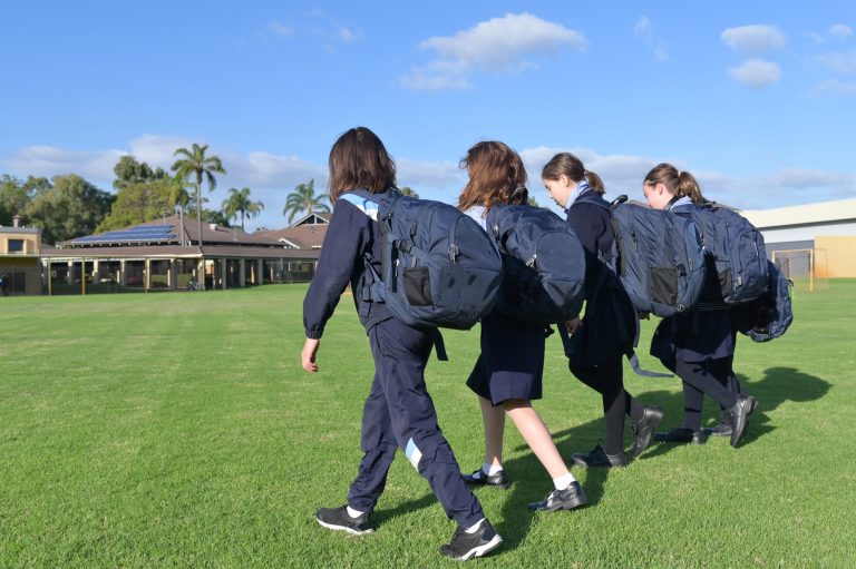 Four students in school uniform with backpacks walking across a school oval in Bendigo, Victoria on a sunny day.