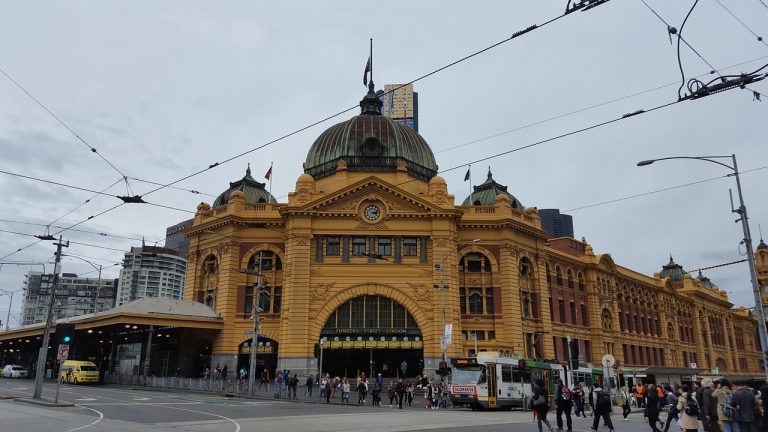 Flinders Street Station with trams and commuters on a cloudy day in Melbourne, Victoria.